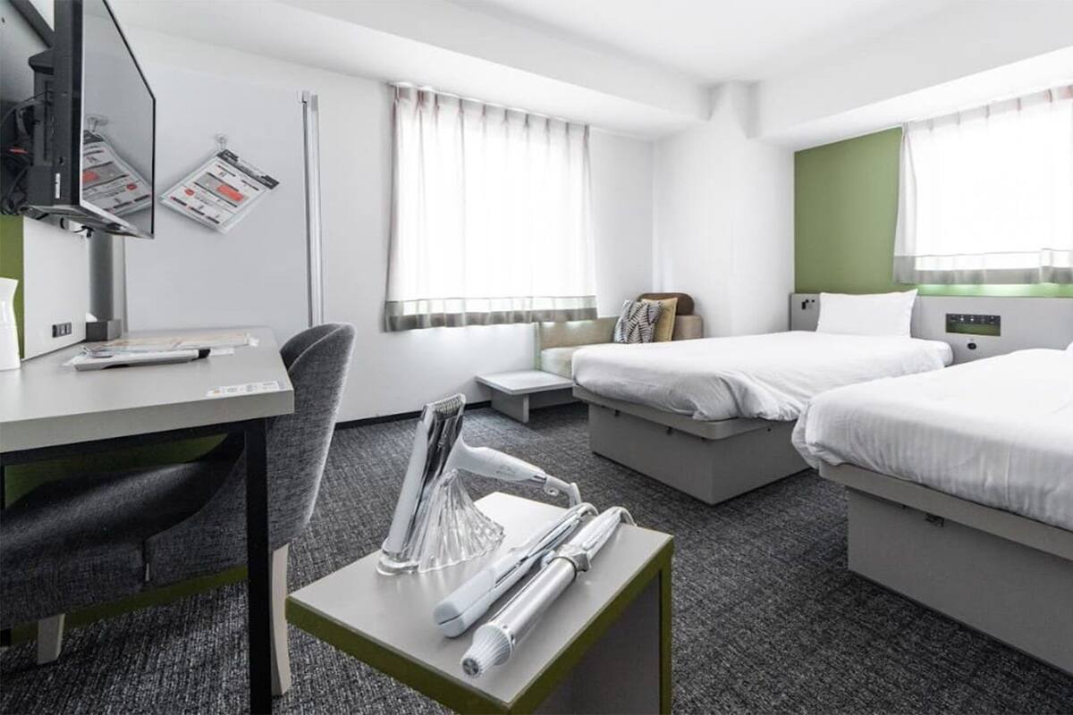 A hotel room with a desk, two beds, a chair, a small table with a telephone and a glass, and a window with curtains.