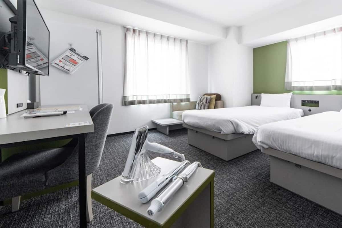 A hotel room with a desk, two beds, a chair, a small table with a telephone and a glass, and a window with curtains.