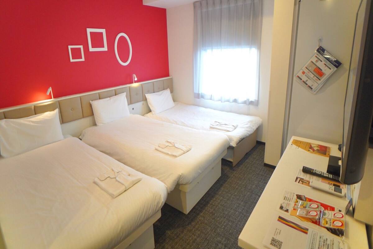 A hotel room with two beds, a red accent wall, a window with curtains, and a desk with a remote control and brochures.