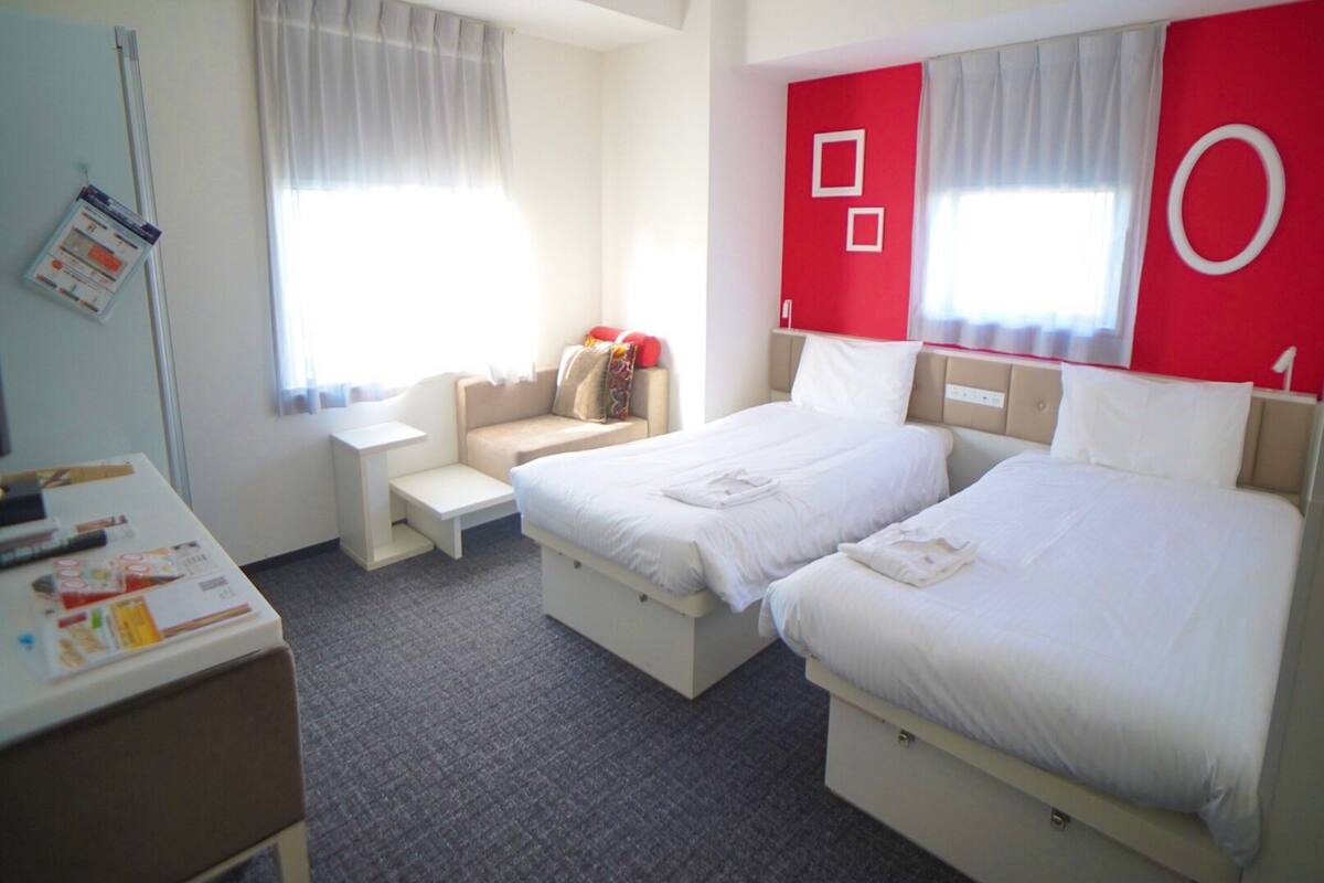 A hotel room with two beds, a small sofa, a desk, and a window with curtains.