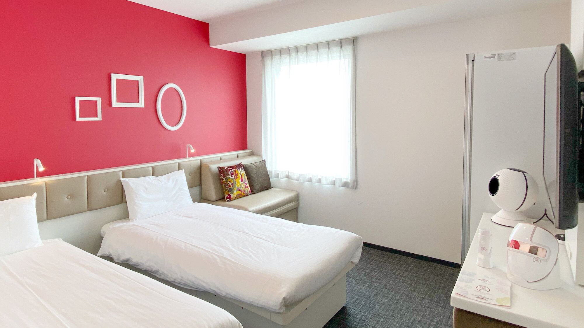 Hotellihuone, jossa on kaksi sänkyä, punainen tehosteseinä, valkoisilla verhoilla varustettu ikkuna, yöpöytä lampulla ja minibaari.