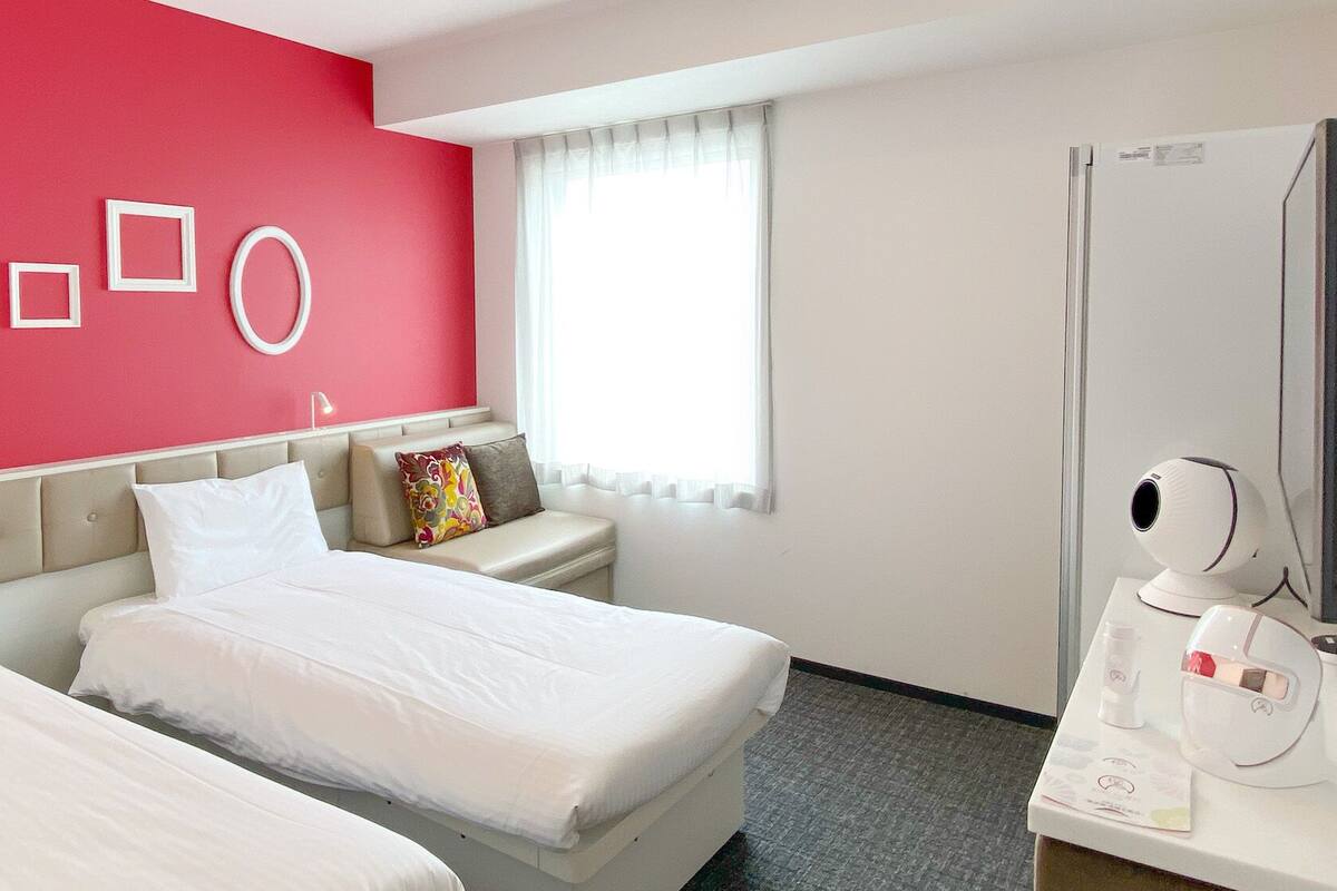 A hotel room with two beds, a red accent wall, a window with white curtains, a bedside table with a lamp, and a minibar.