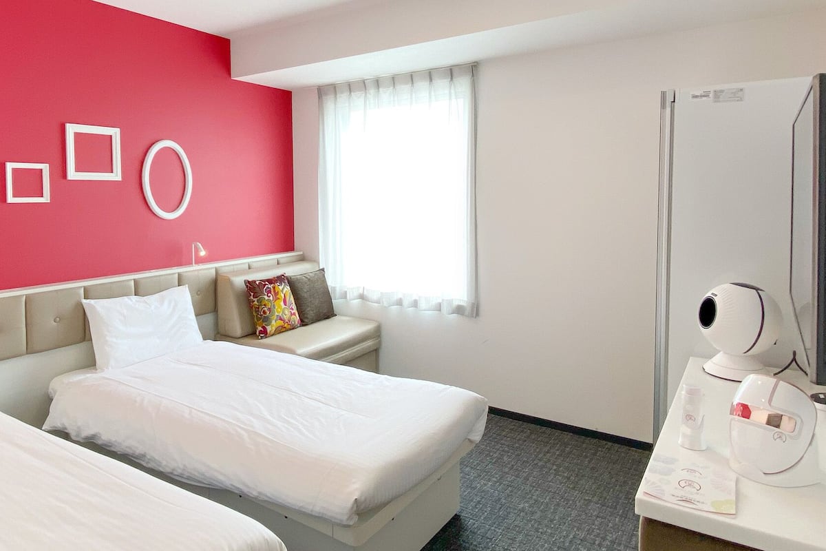 A hotel room with two beds, a red accent wall, a window with white curtains, a bedside table with a lamp, and a minibar.