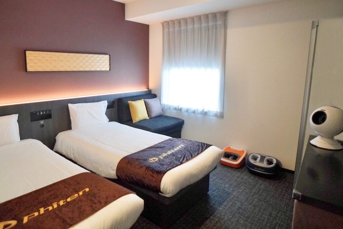 A hotel room with two beds, a desk, a chair, a window with curtains, a webcam, and a shoe rack.