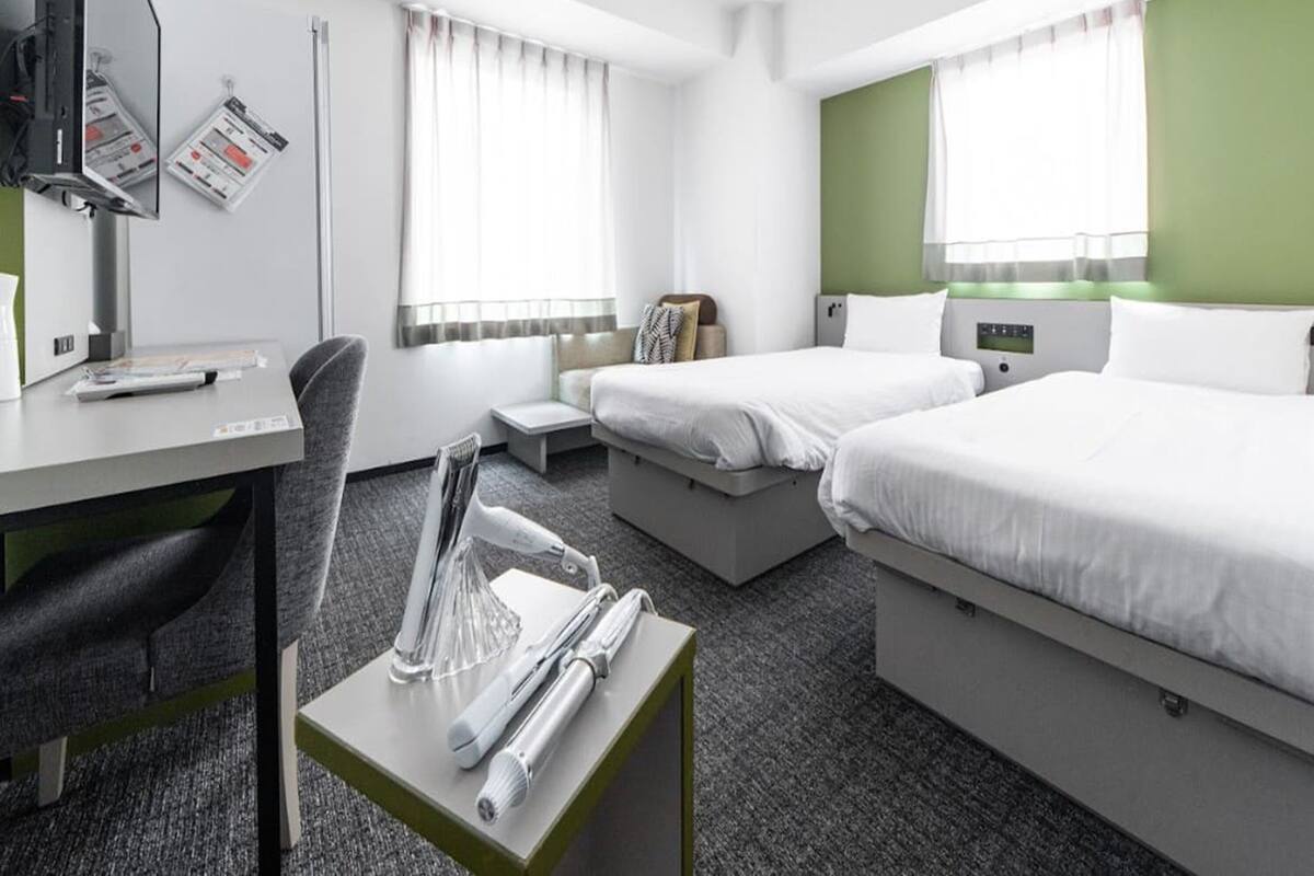 A hotel room with two beds, a desk, a chair, a small table with toiletries, and a window with curtains.