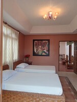 A room with two beds, a painting on the wall, a wooden headboard, a vase with flowers, and a chandelier.