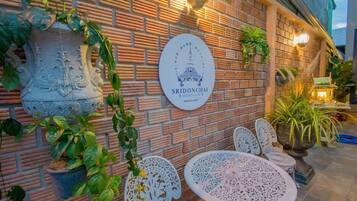 A brick wall with a sign reading "SRIDONCHAI HERITAGE" and decorative metalwork. There are white metal chairs and a table, and potted plants are visible.