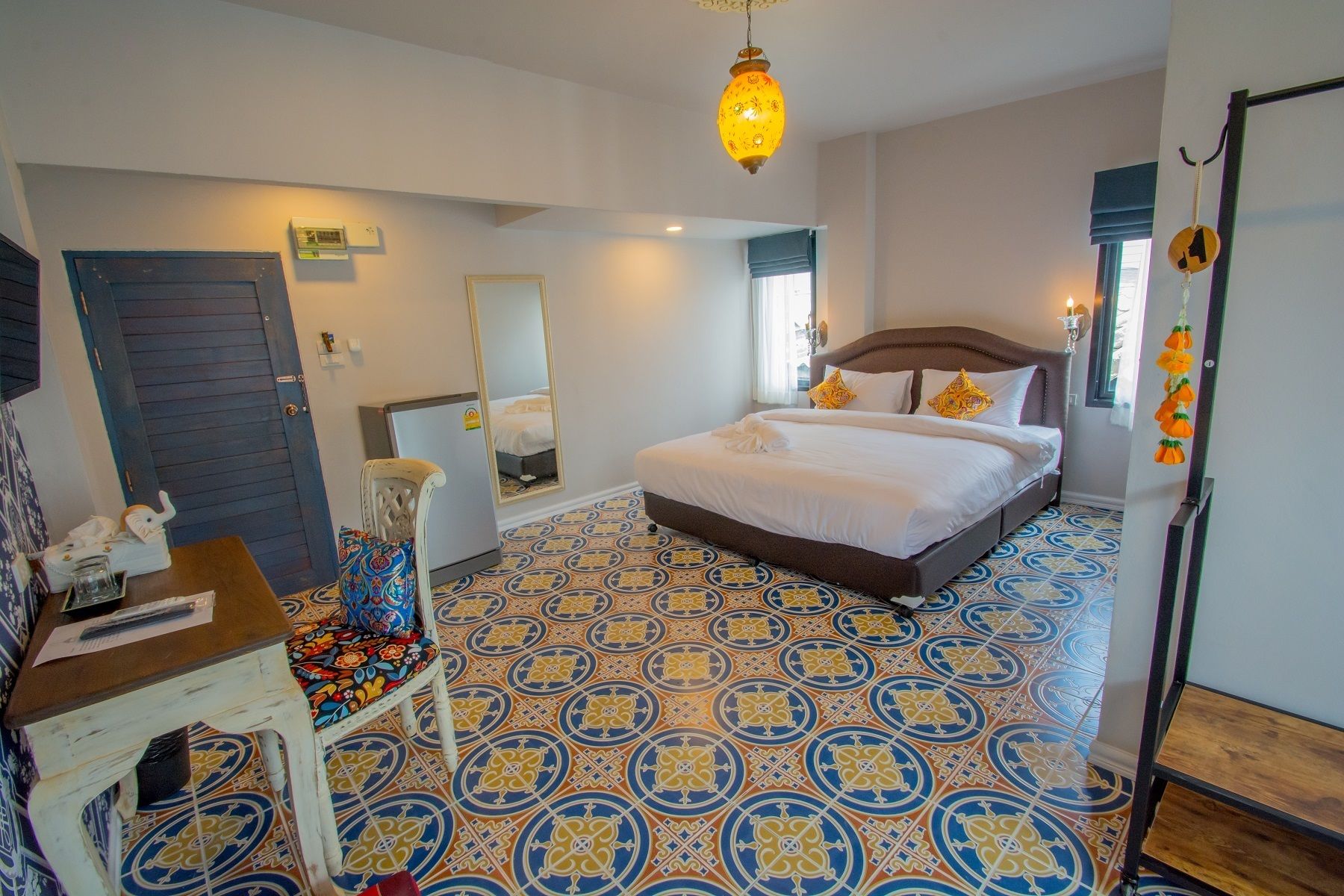 A bedroom with a patterned floor, a bed with white bedding, a mirror, a desk with a chair, and a hanging light fixture.