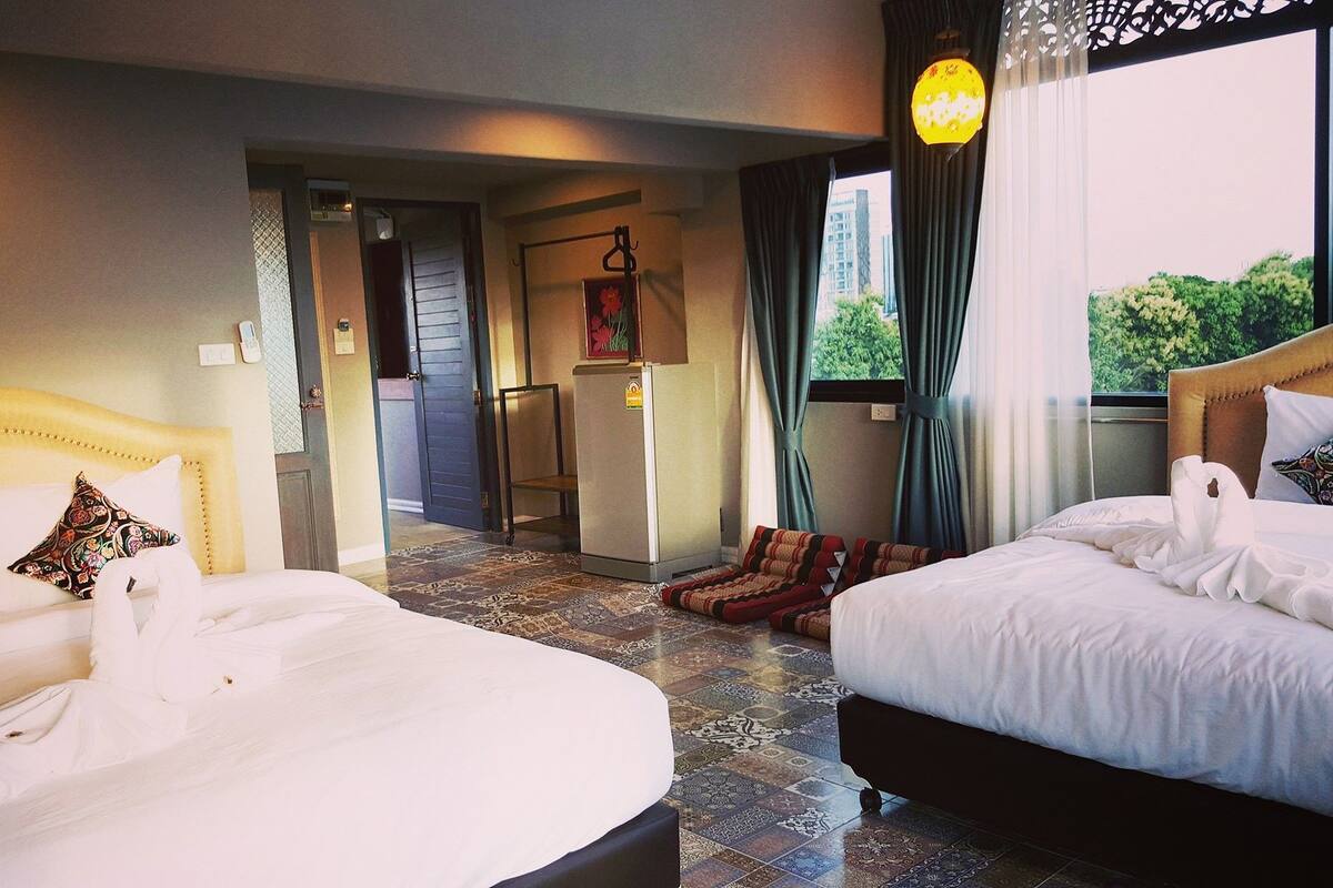 Ein Hotelzimmer mit zwei Betten, einem Kühlschrank, einer Tür zu einem anderen Raum und einem Fenster mit Vorhängen.