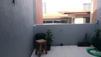 A small, enclosed balcony with tiled flooring, a wooden stool, a black trash bin, and potted plants.