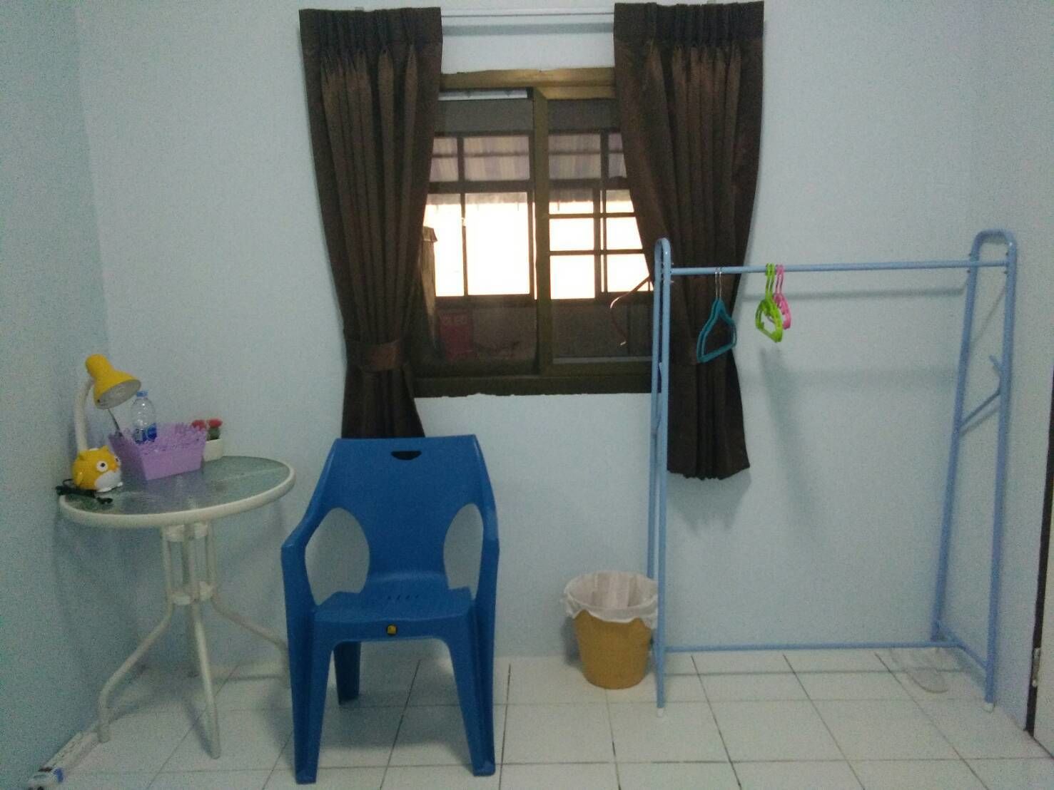 A small room with a window, a blue plastic chair, a small table with a lamp, and a clothes drying rack.