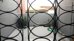 A view through a metal gate with a circular pattern, leading to a courtyard with a tiled floor, a green hose, and some potted plants. - A & P House Charansanitwong (Bangkok)