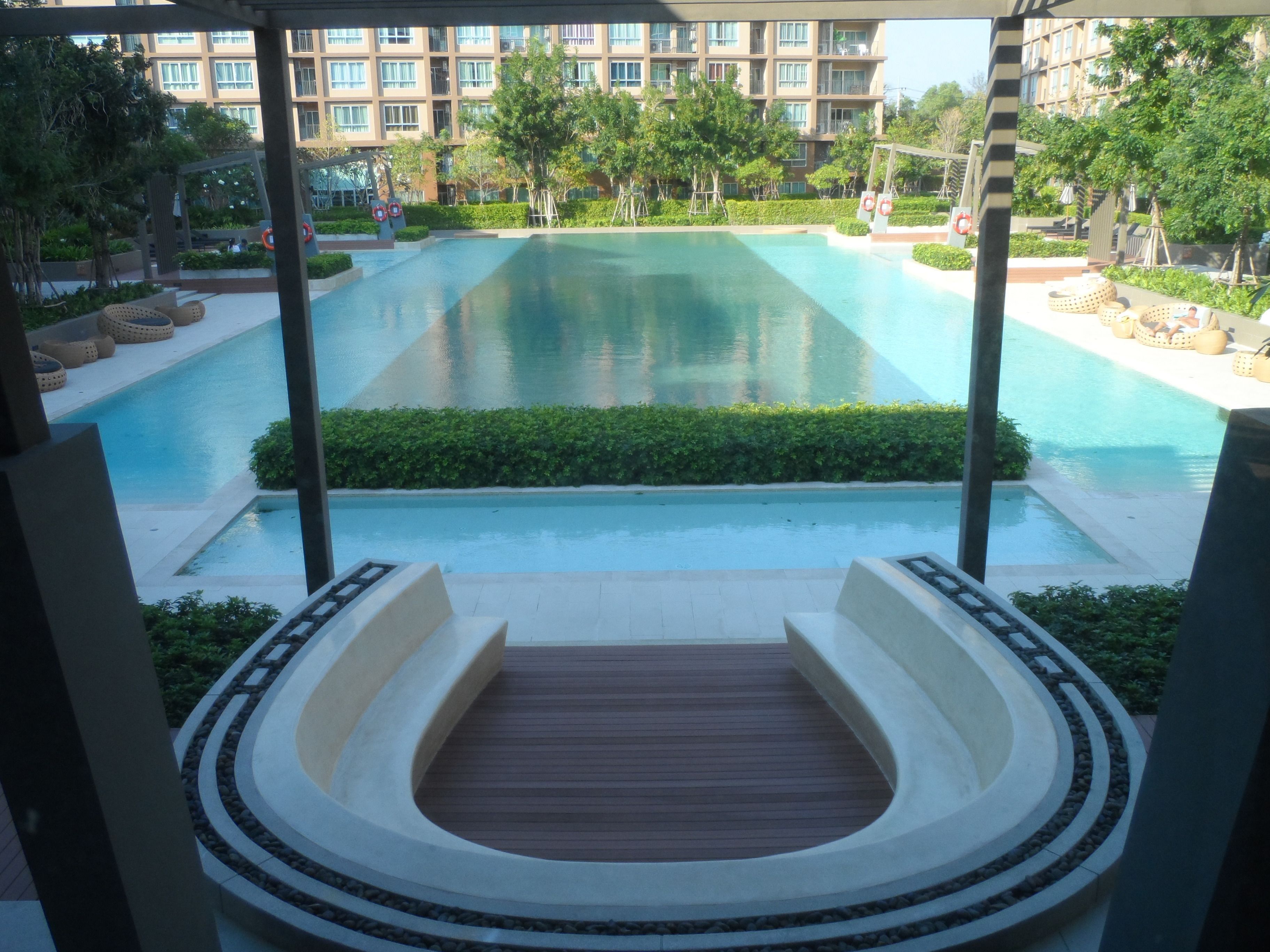 A large rectangular pool with a wooden deck in the foreground, surrounded by greenery and a multi-story building in the background.