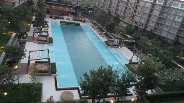 A large rectangular swimming pool surrounded by greenery and modern buildings. - Baan Thew Lom By Woranan (Cha-am)