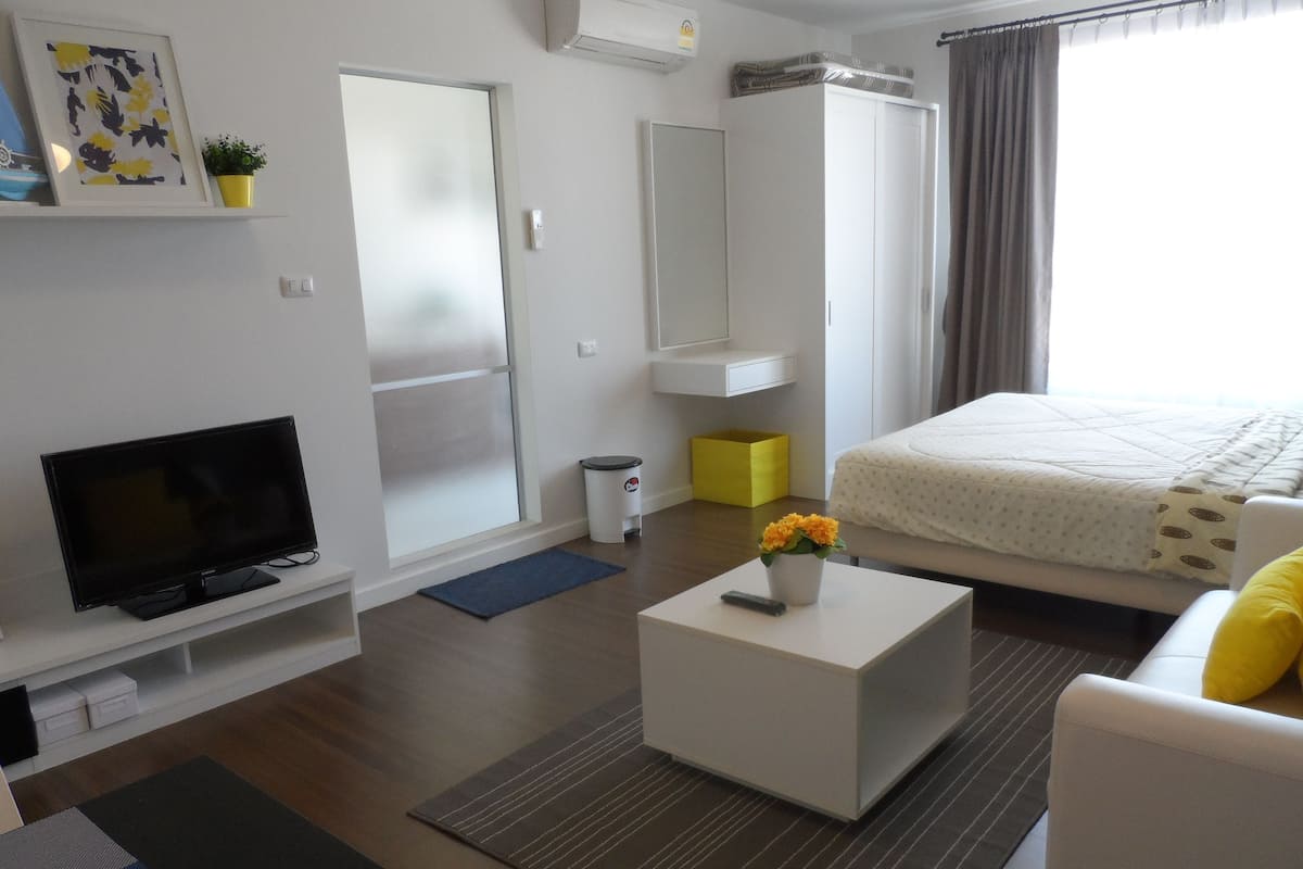 A modern hotel room with a flat-screen TV, a bed with a white duvet, a small white table with a vase of flowers, and a large window with curtains.
