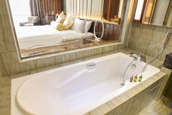 Un baño moderno con una bañera grande, un tocador con mampara de vidrio y un espejo que refleja un dormitorio con cama y cortinas.