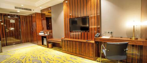 A hotel room with a wooden panel wall, a flat-screen TV mounted above a desk, a chair, and a patterned carpet.