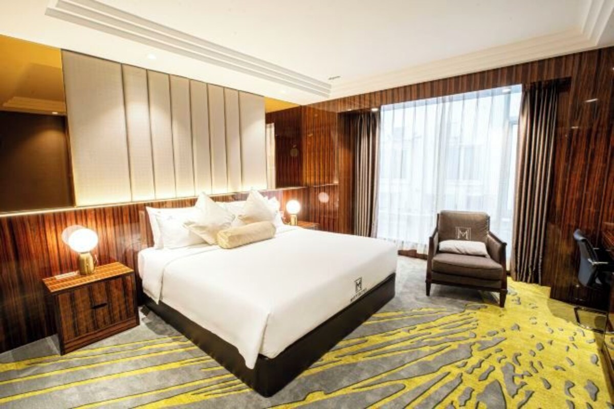 Habitación de hotel con una cama grande, una mesita de noche con lámpara, una silla y un ventanal con cortinas.