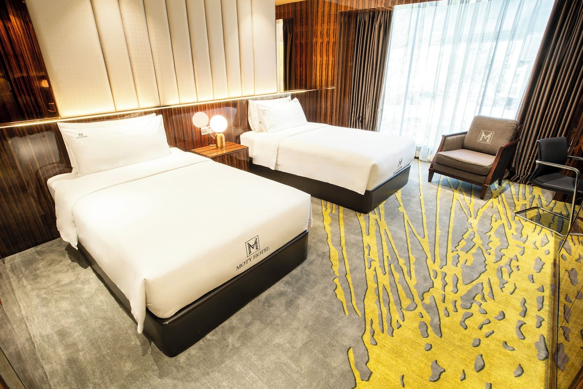 Une chambre d’hôtel avec deux lits, un coin salon avec un fauteuil et une grande fenêtre avec des rideaux.