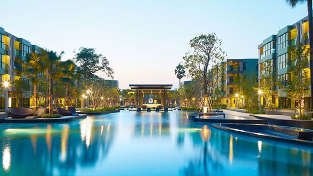 Zona de piscina de hotel con un edificio central, rodeada de edificios modernos y palmeras.