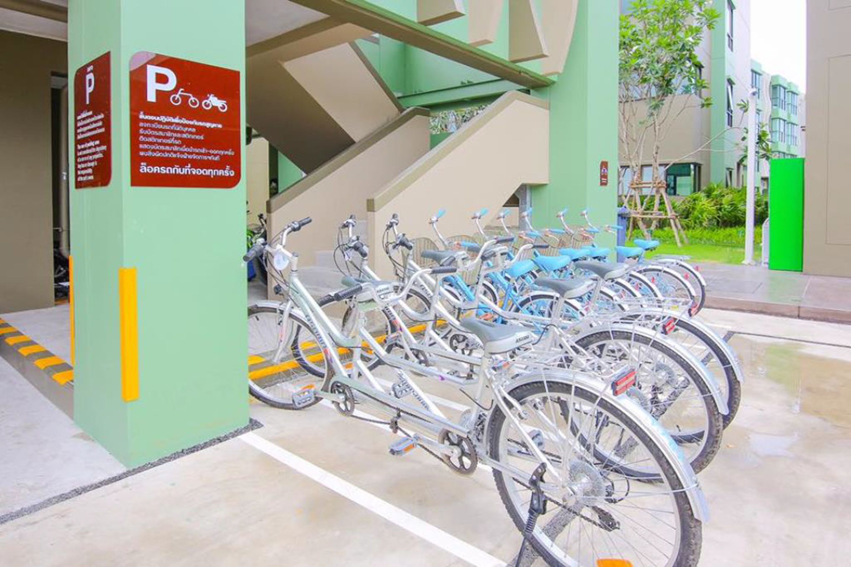 A bike parking area with multiple bicycles parked in rows, a sign with parking information, and a staircase in the background.