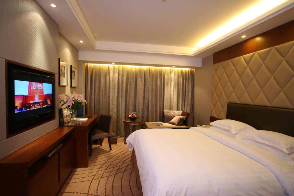Een hotelkamer met een groot bed, een tv aan de muur, een bureau met een stoel en een kleine tafel met een bloevas.