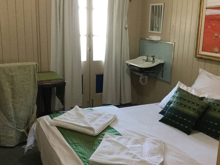 Habitación de hotel con cama, colcha verde, dos toallas blancas, almohada verde, una mesita con silla, ventana con cortinas y un lavamanos instalado en la pared.