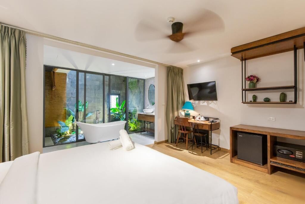 A hotel room with a large bed, a desk with a chair, a television, a small refrigerator, and a sliding glass door leading to a balcony with a bathtub and greenery.