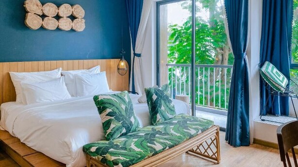 Quarto com cama, sofá, cabeceira de madeira, janela com cortinas e vista para o verde lá fora.
