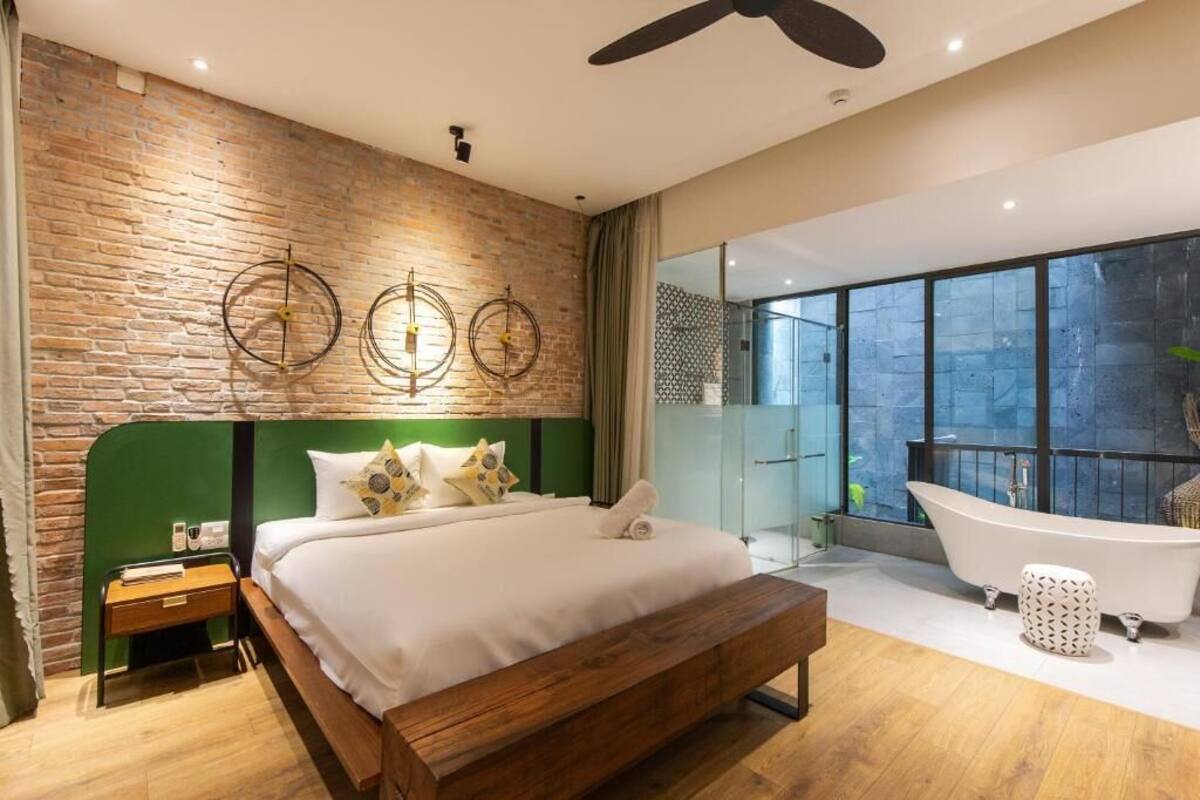 Une chambre à coucher moderne avec un mur en briques, un grand lit, une tête de lit verte, un banc en bois, une table de chevet et une grande fenêtre, ainsi qu’une baignoire sur pieds.