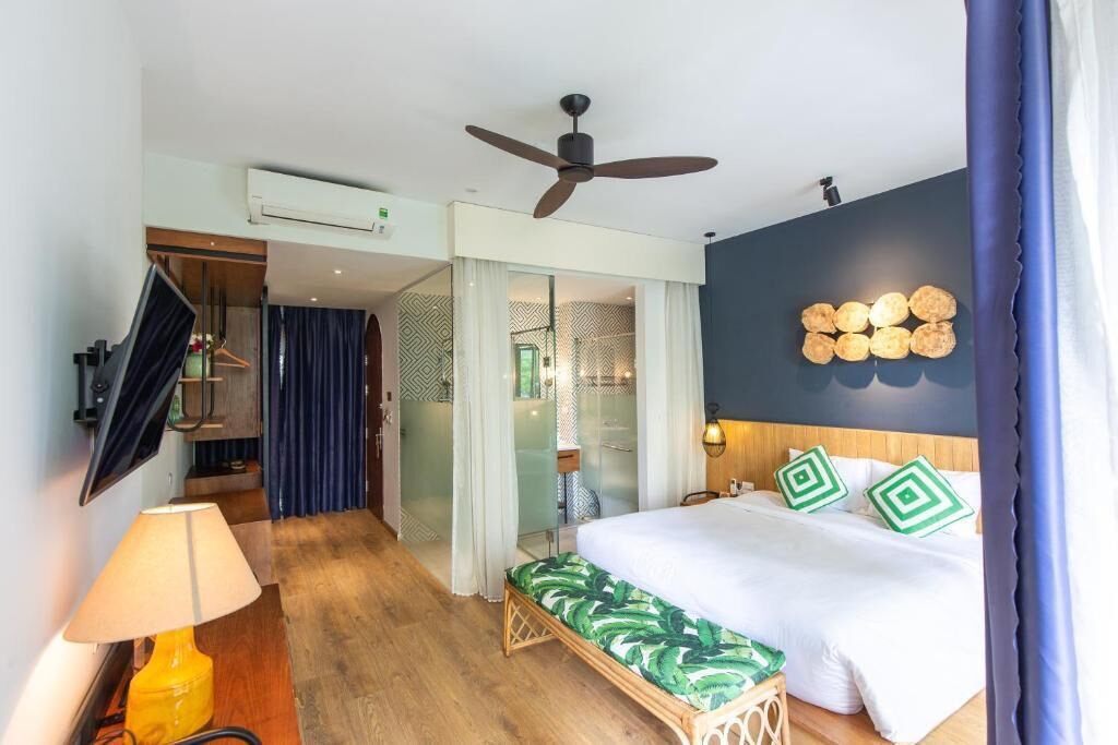 A hotel room with a bed, a ceiling fan, a wall-mounted TV, and a bathroom visible through a glass door.
