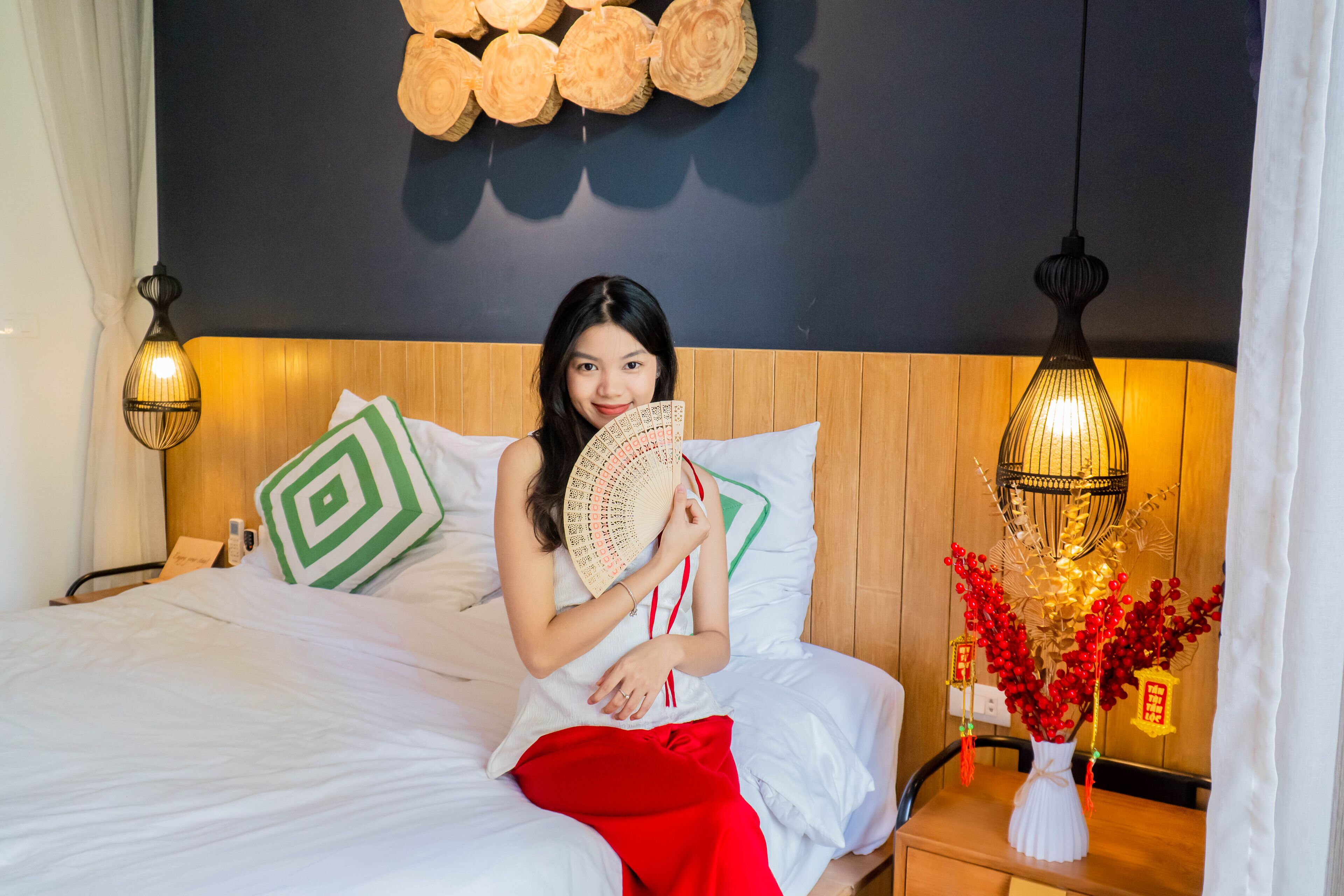 A bed with white bedding, a green and white pillow, and a red blanket. There is a bedside table with a white vase and red flowers.
