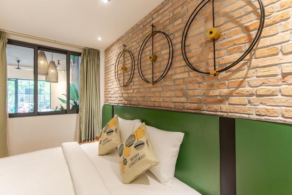 A hotel room with a brick wall, a green headboard, and a bed with white bedding and Christmas-themed pillows.