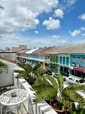 A balcony with potted plants, a white table, and a view of a street with buildings and cars. - Xinlor House (Phuket)
