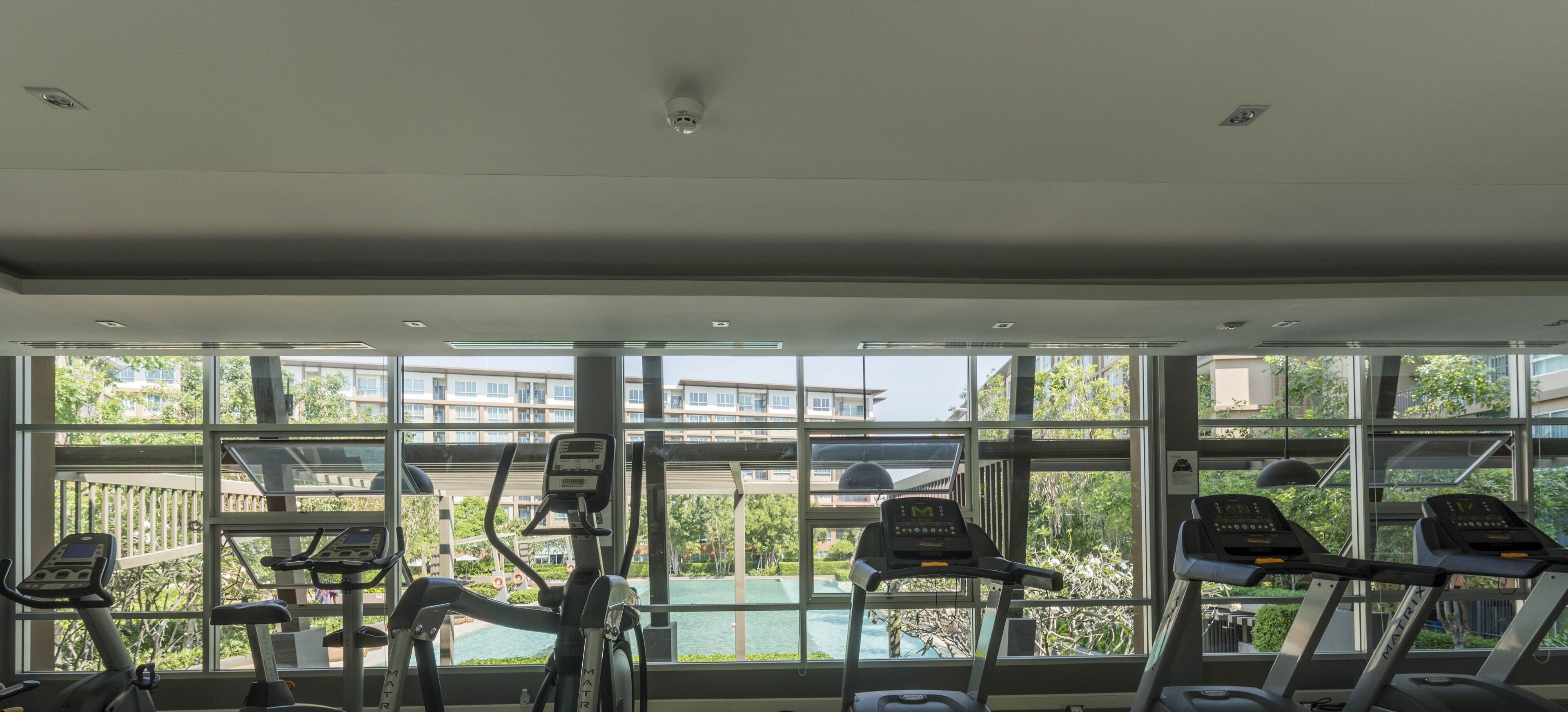 Un gimnasio con cintas de correr y bicicletas estáticas, ventanas amplias con vistas a edificios y zonas verdes.