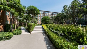 A pathway flanked by lush greenery and a modern building with multiple windows. - Baan Thew Lom Cha-Am Huahin by D-light (Cha-am)