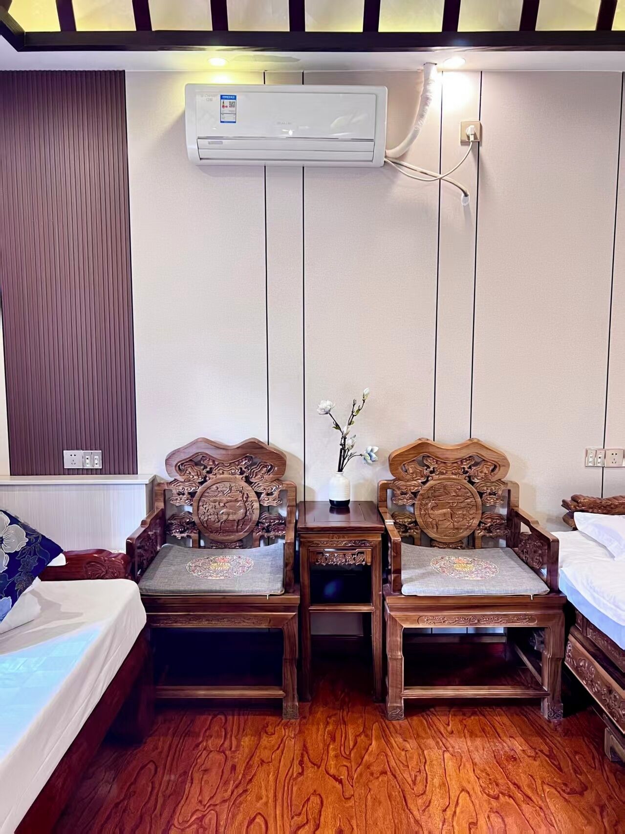 A traditional room with a bed, two wooden chairs with intricate carvings, a small table with a vase, and an air conditioning unit mounted on the wall.