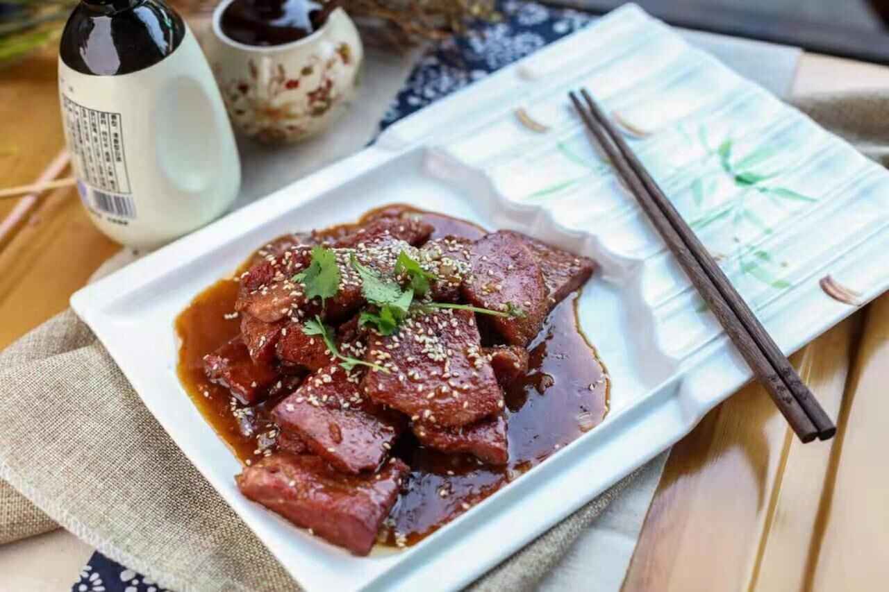 Een bord vlees met een donkere saus, versierd met sesamzaadjes en koriander, met stokjes en een kleine schaal saus.
