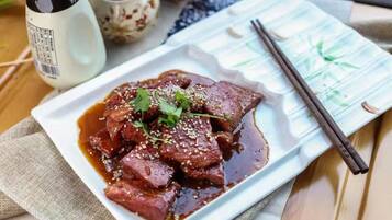 Een bord vlees met een donkere saus, versierd met sesamzaadjes en koriander, met stokjes en een kleine schaal saus.