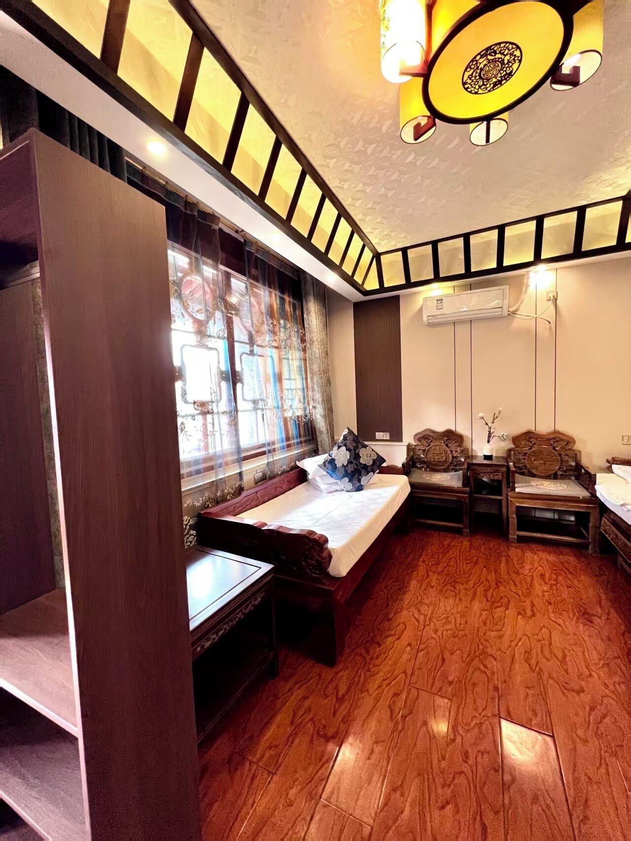 A bedroom with a bed, wooden floor, a window with curtains, a nightstand, and a wall-mounted air conditioner.
