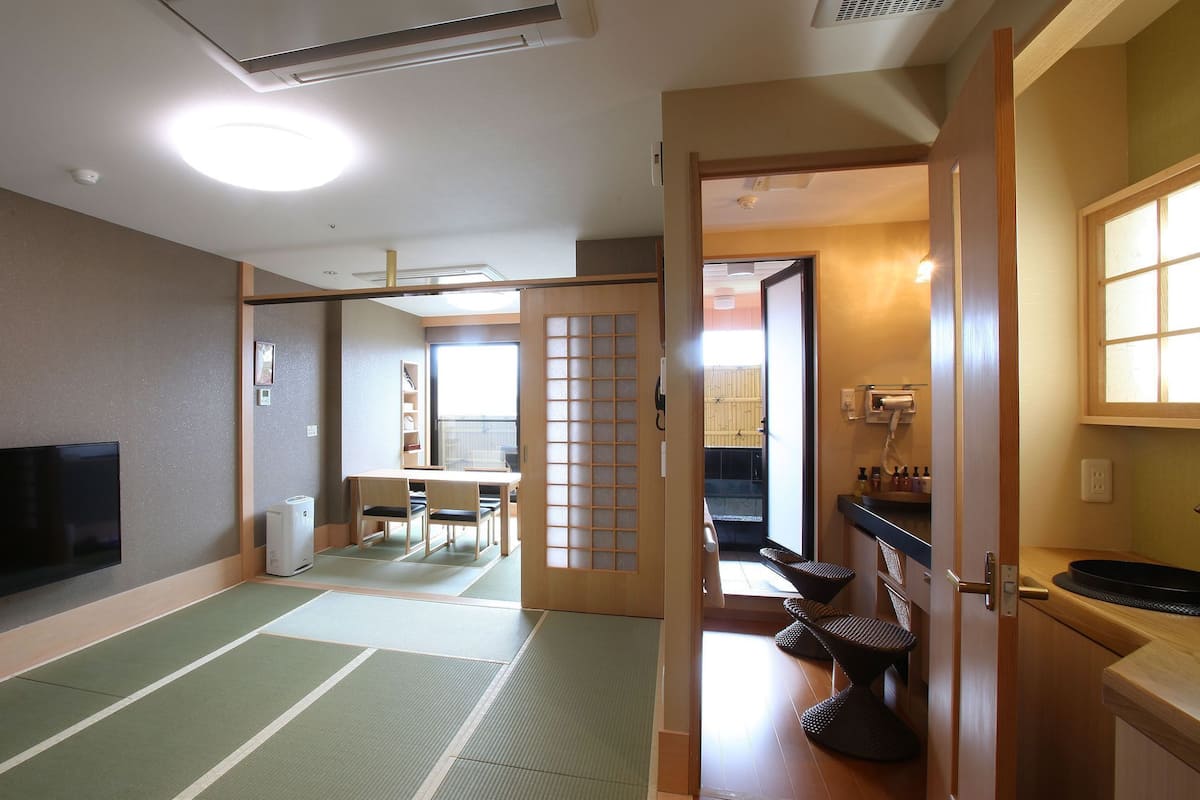 A traditional Japanese room with tatami flooring, a television, a sliding door leading to a balcony, and a small dining area.