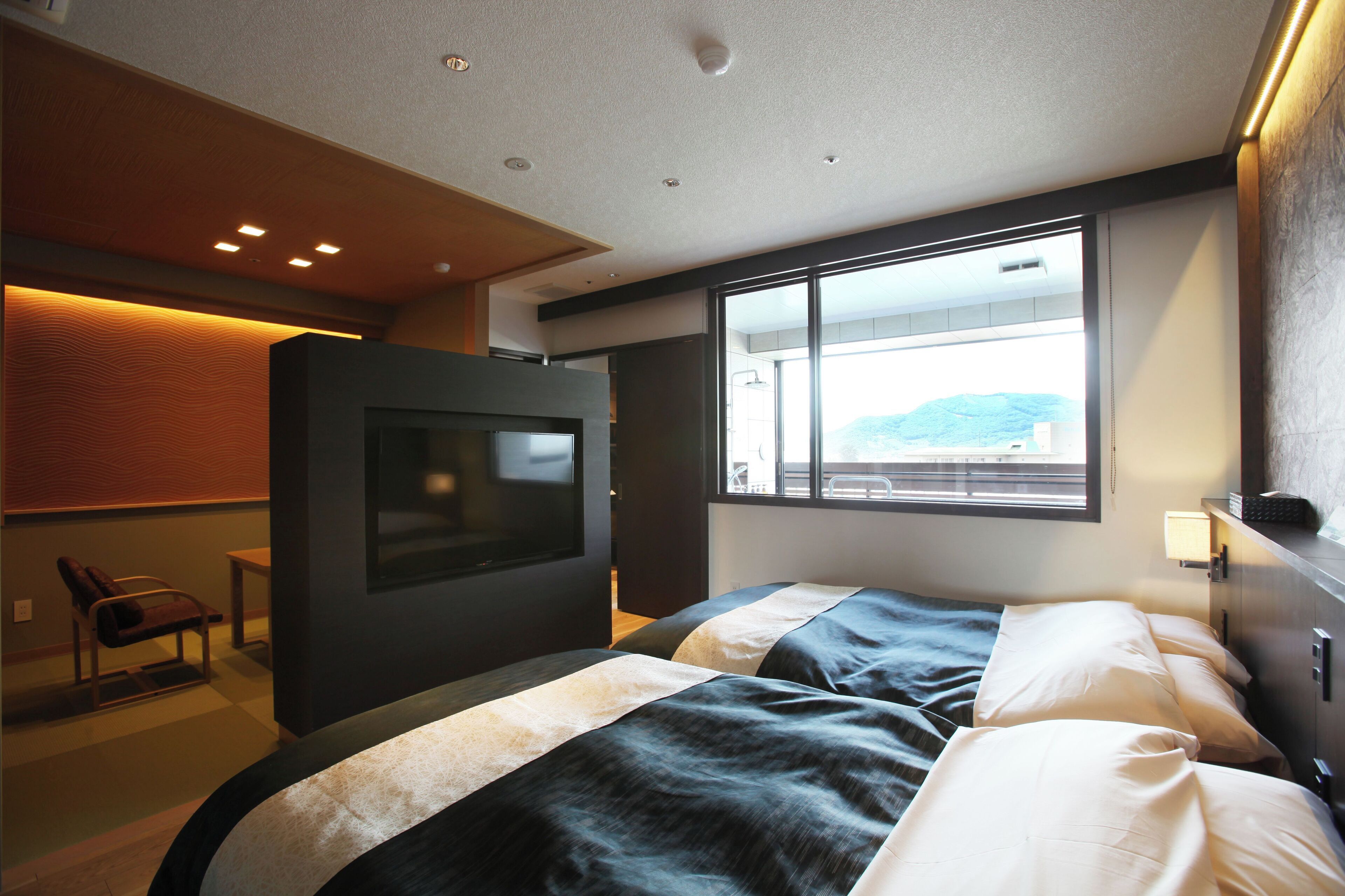 A modern hotel room with a large bed, a television, and a view of the outdoors through a large window.