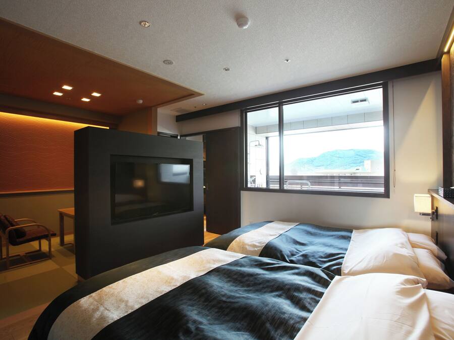 A modern hotel room with a large bed, a television, and a view of the outdoors through a large window.