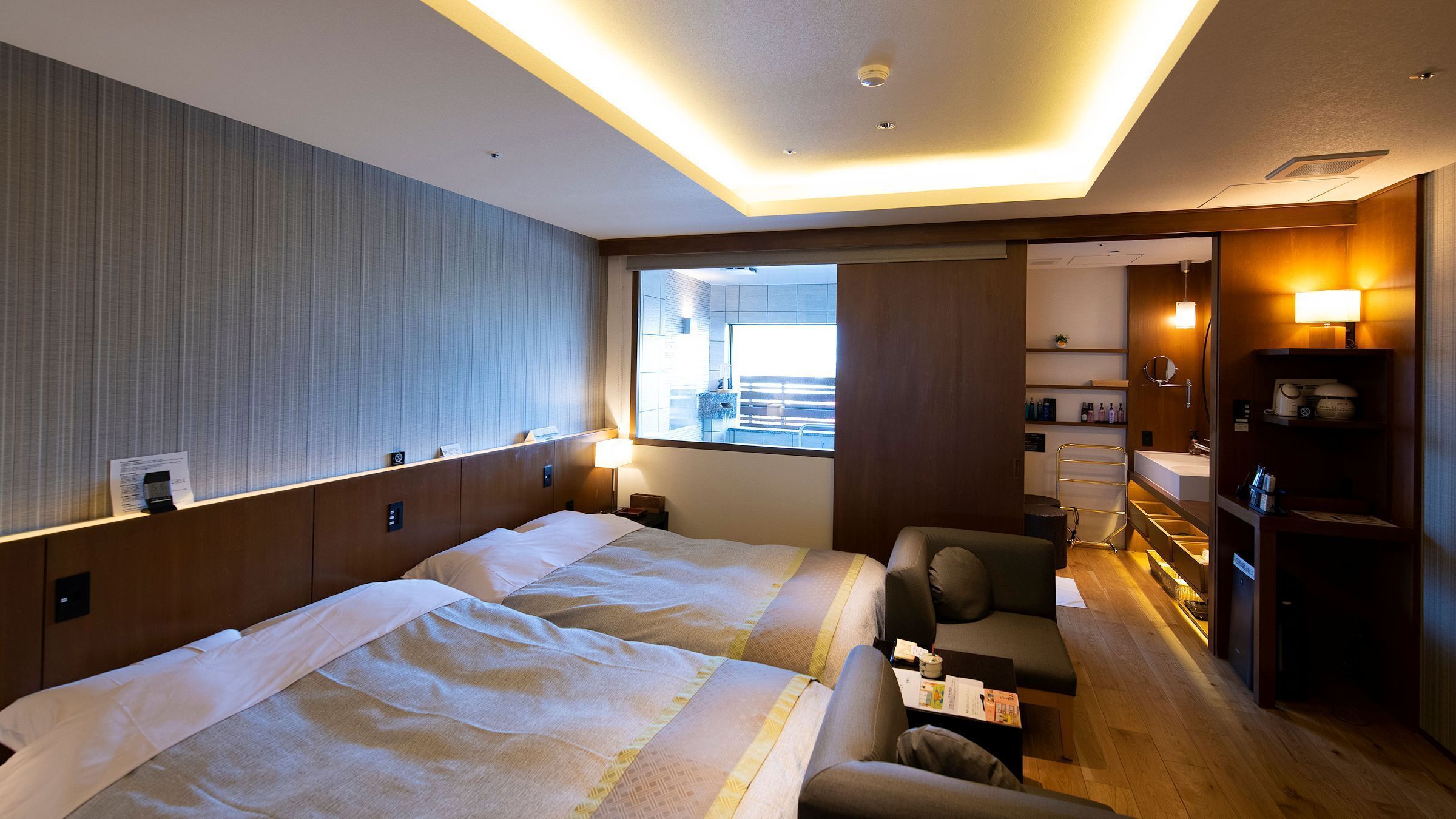 A hotel room with a large bed, a sofa, a small table, a desk, a mirror, a window, and a bathroom with a sink and shelves.