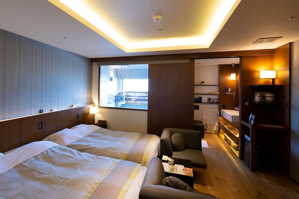 A hotel room with a large bed, a sofa, a small table, a desk, a mirror, a window, and a bathroom with a sink and shelves.