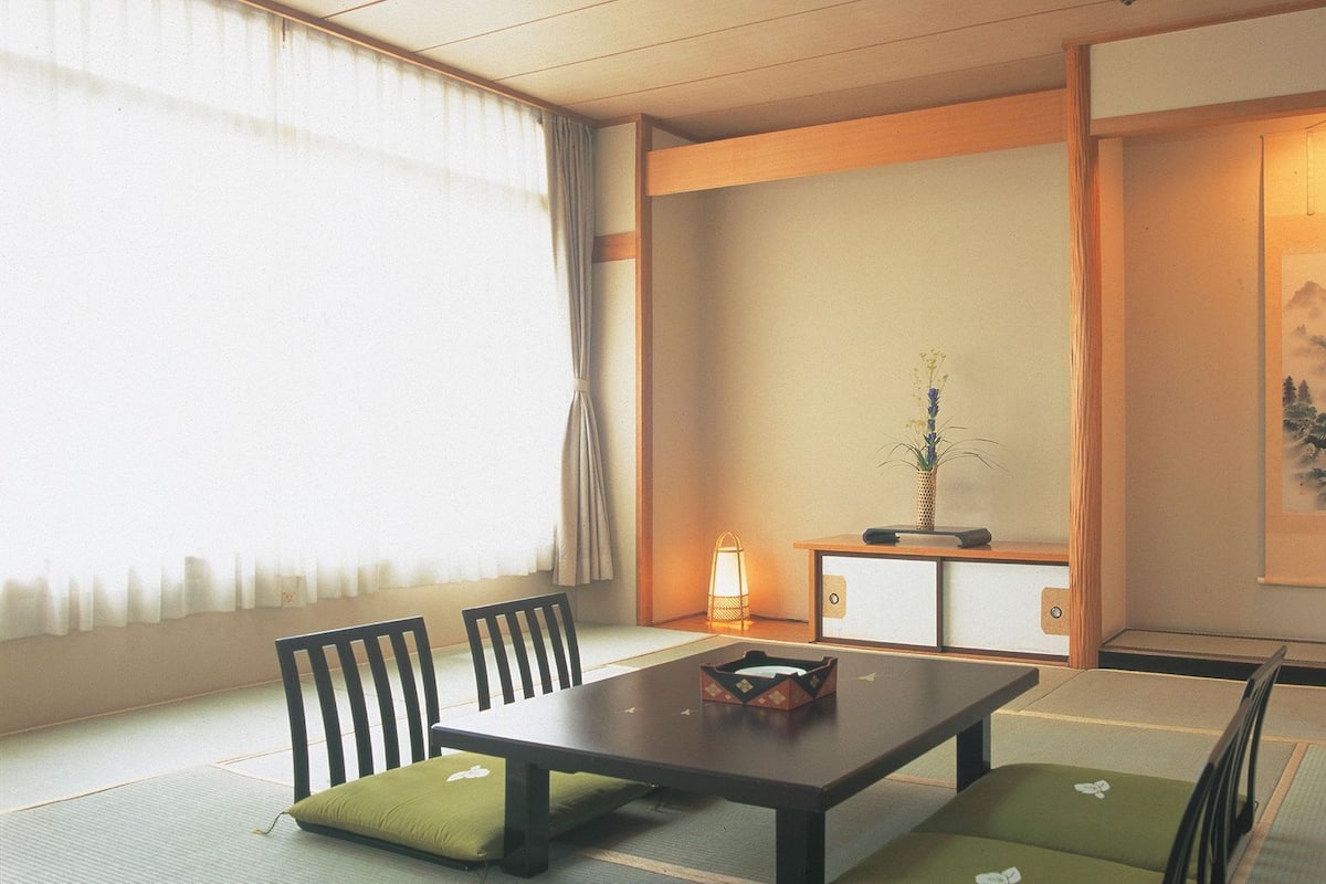 A traditional Japanese room with tatami flooring, a low table, chairs, a small table with a vase, and a painting on the wall.