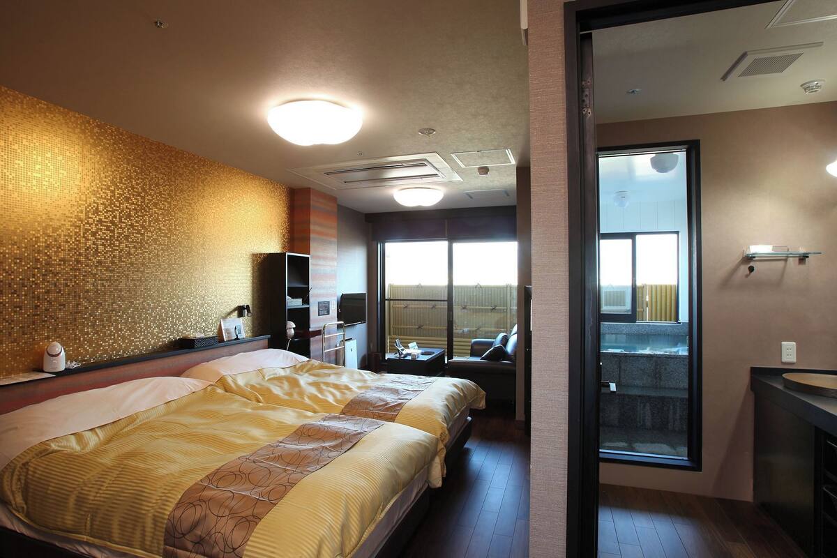A hotel room with a bed, a desk, a chair, a large window, and a bathroom with a mirror and sink.