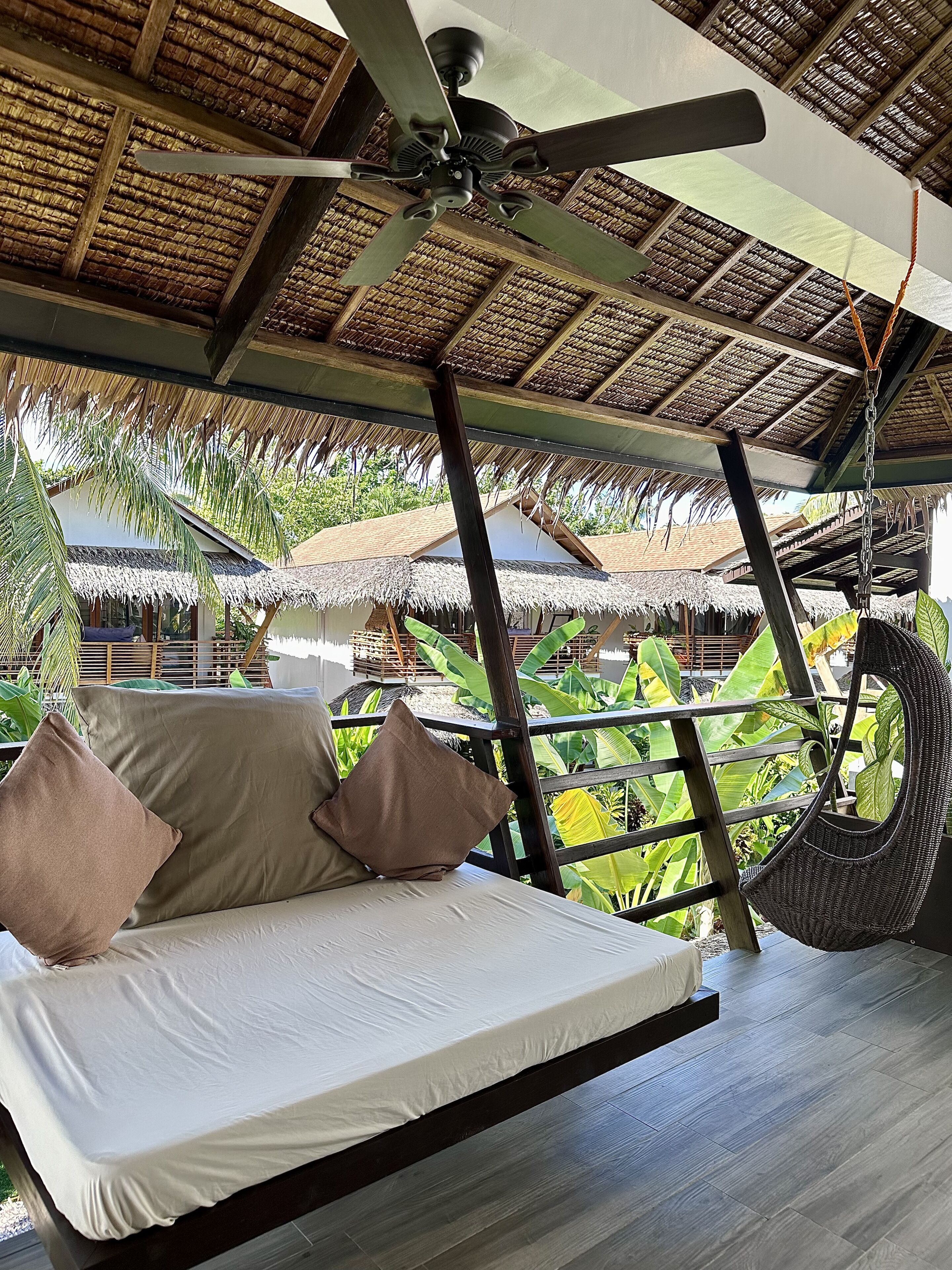 A bed with pillows, a ceiling fan, and a view of a thatched-roof structure and lush greenery.