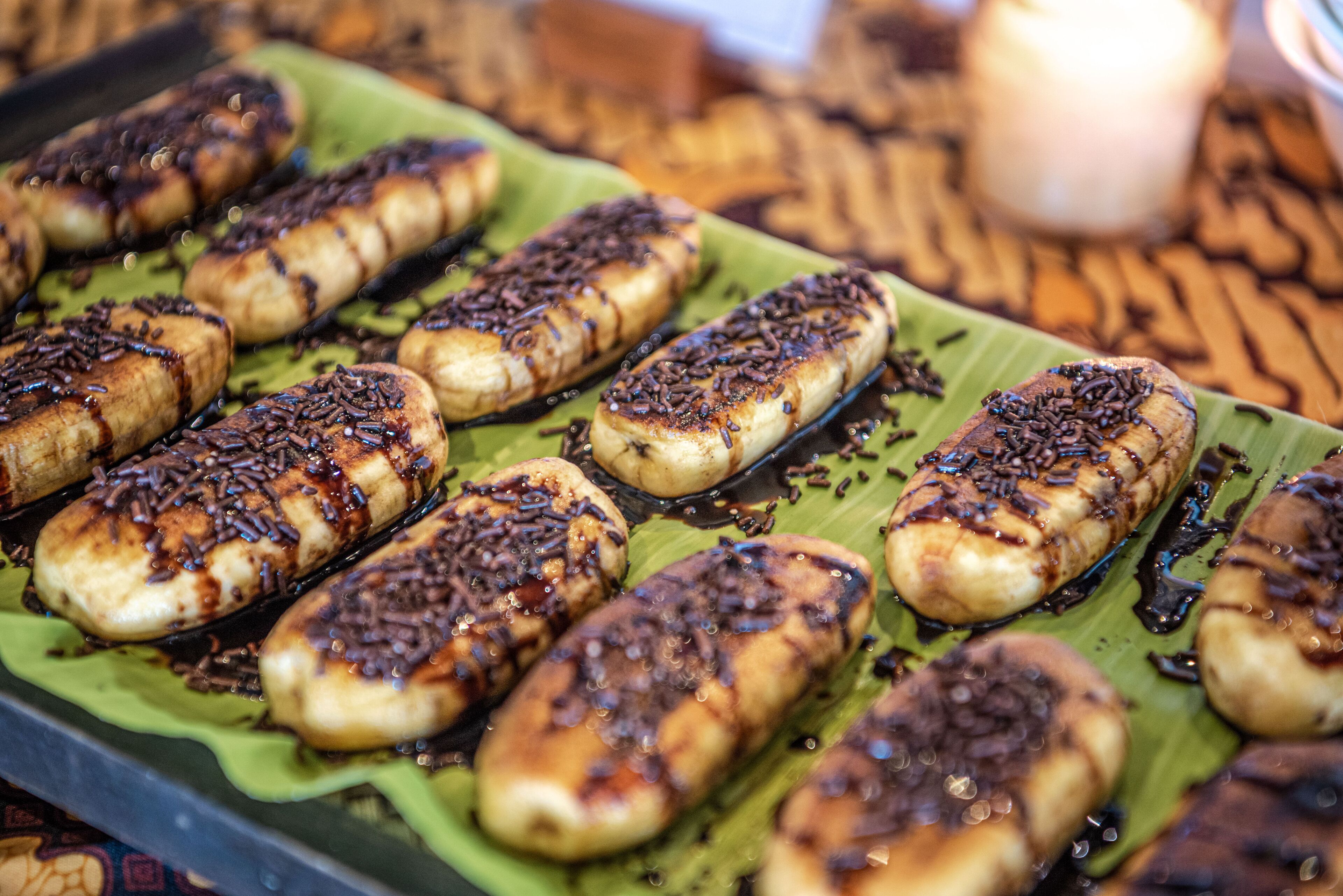 A tray of grilled bananas with chocolate drizzle on a green leaf.