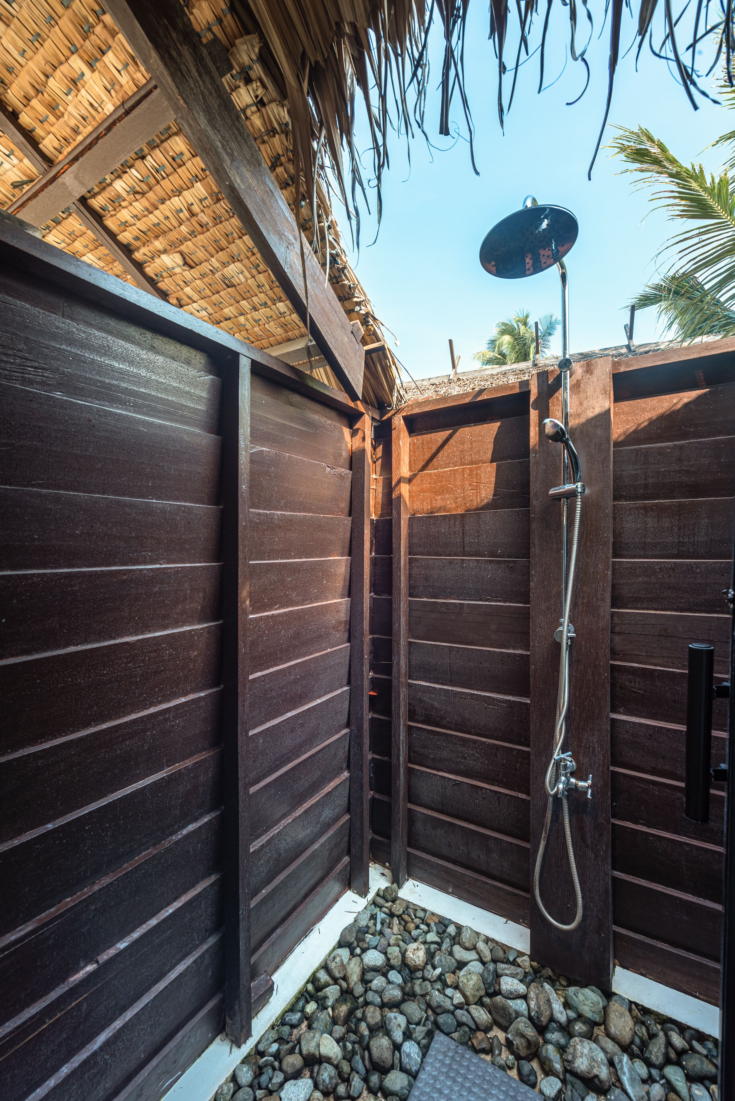 A wooden shower enclosure with a metal showerhead and hose, surrounded by a thatched roof and palm trees.